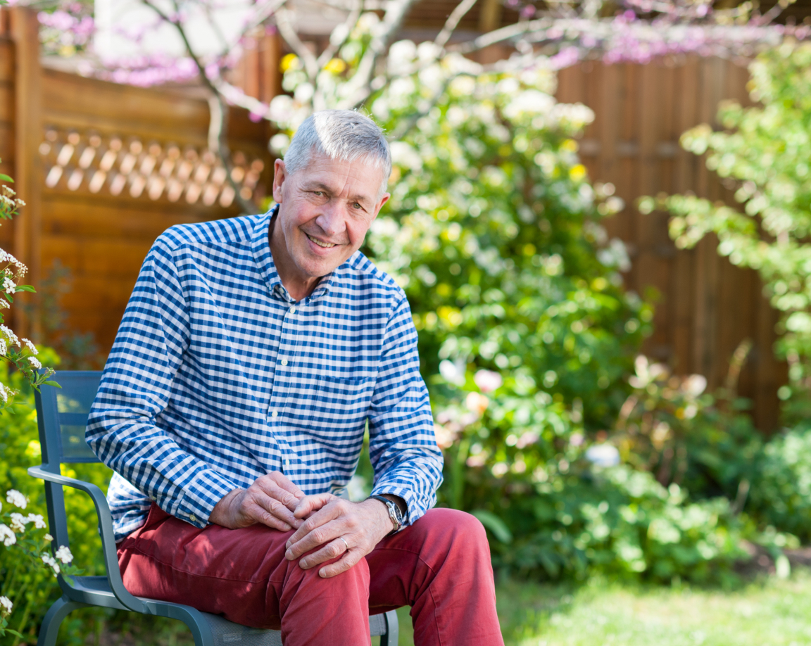 A senior man smiling to the camera while sitting on a chair in front of a nice back yard