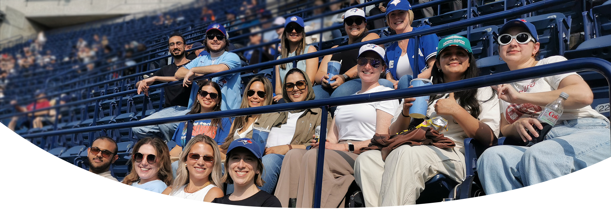 15 people who work together sitting in a baseball stadium with baseball caps and sunglasses smiling for a picture.